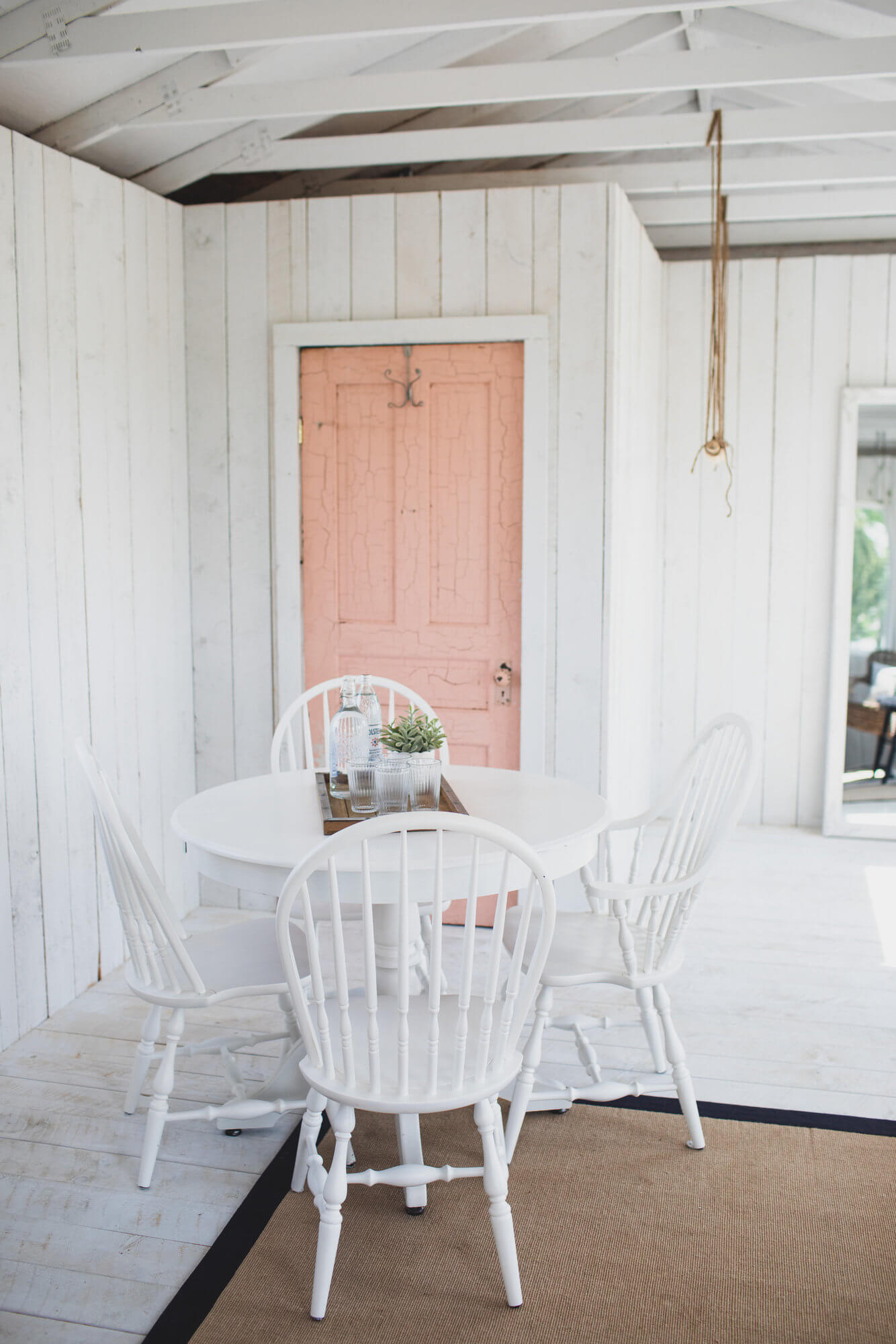 garden cottage white shiplap walls with a white roof and a blush pink door, in the room is a white table and 4 white chairs a beige rug and water glasses sitting on the table